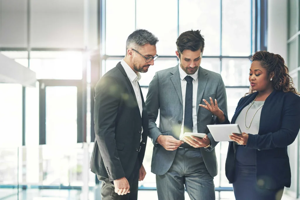 A diverse group of three professional business colleagues in formal suits standing in a bright, modern office lobby, discussing work while looking at a digital tablet.