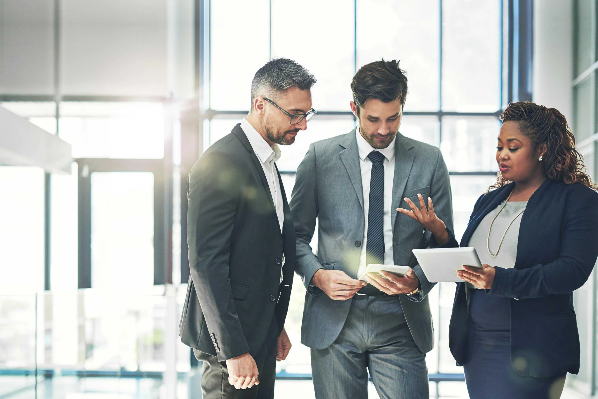 A diverse group of three professional business colleagues in formal suits standing in a bright, modern office lobby, discussing work while looking at a digital tablet.