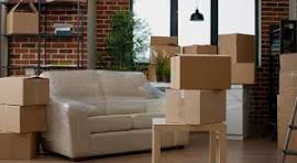 A modern living room with a white sofa and a brick wall, filled with numerous stacked cardboard boxes ready for a move.