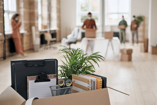 A close-up of an open cardboard box filled with office supplies and a small plant, with a blurred background showing people moving in a bright office.