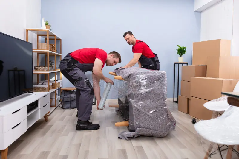Two professional movers in red and grey uniforms carefully wrapping a grey sofa with plastic stretch film in a modern living room.