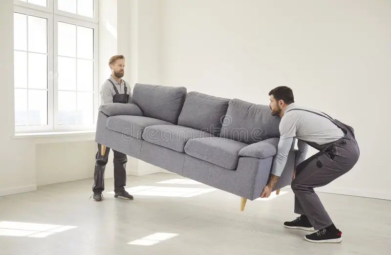 Two professional movers in grey uniforms lifting and carrying a large grey sofa across an empty, bright room.